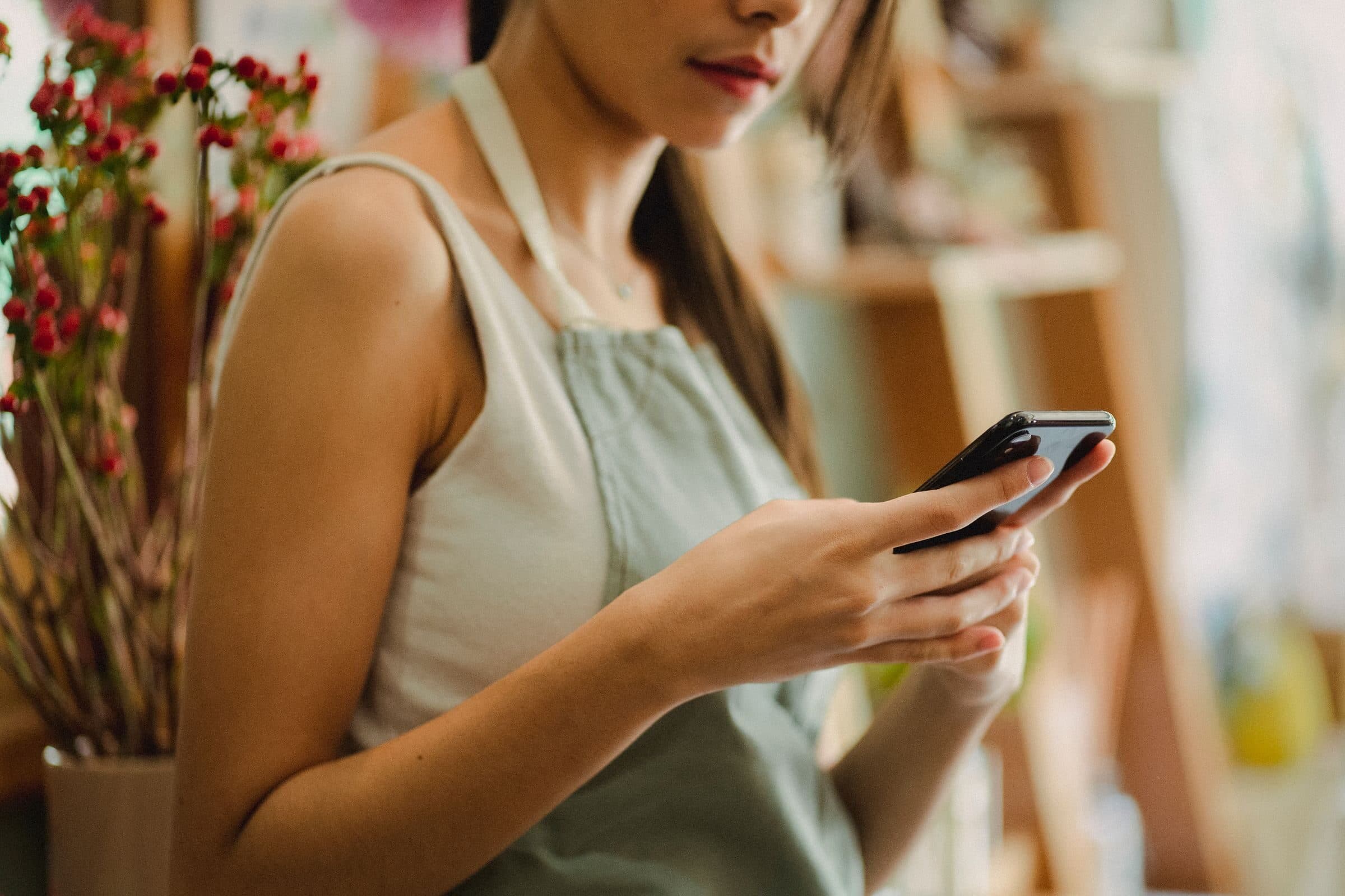A shop worker in an apron checking her phone in a store, a small business that needs general liability for customer injuries and property damage