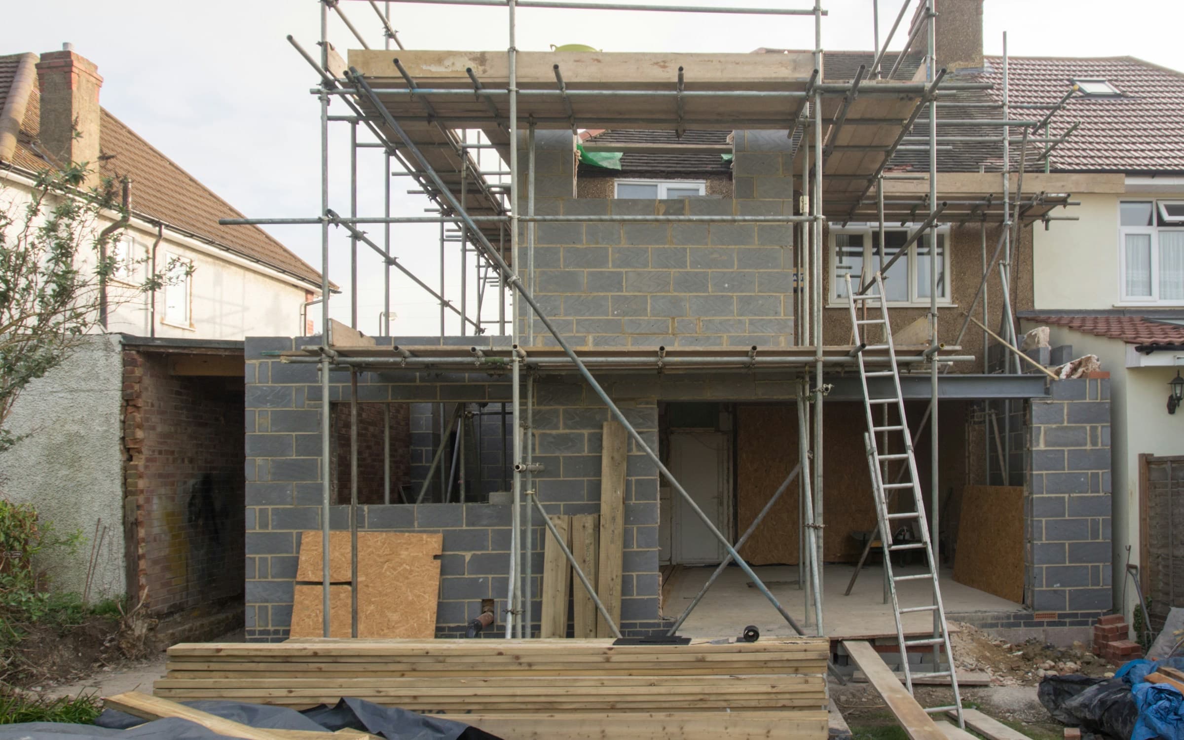 Una ampliación de dos pisos en construcción sobre una casa existente, con muros de bloque dentro de un andamio y madera apilada al frente, la obra que cubre una póliza de renovación